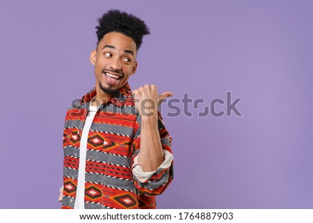 Side view of funny young african american guy in casual colorful shirt posing isolated on violet background studio portrait. People emotions lifestyle concept. Mock up copy space. Point thumb aside