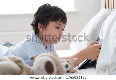 6-7 year old boy playing with dog toy and watching cartoon on tablet,Happy child lying in bed playing game in the morning, Cute Kid having fun and relaxing on his own in bed room,New normal lifestyle 