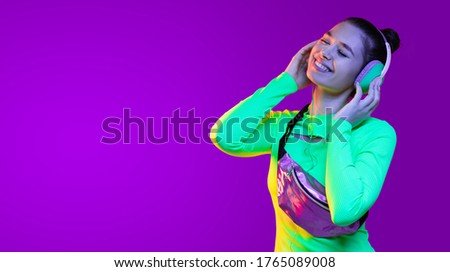 Horizontal banner of young smiling girl dancing in neon light isolated on purple background, enjoying music in headphones, copy space on left