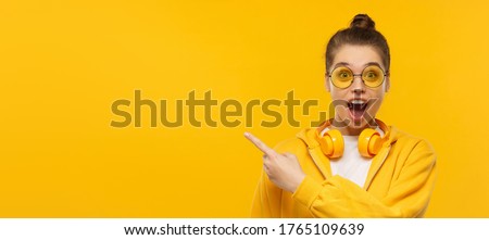 Horizontal banner of young shocked girl, wearing round glasses and headphones on neck, pointing left to copy space, isolated on yellow background