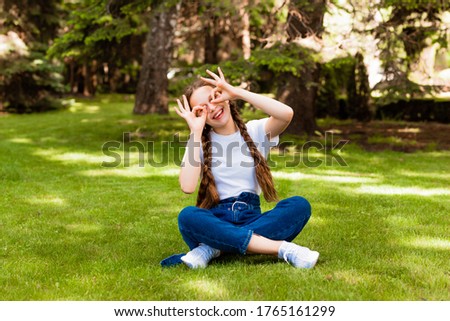 a young girl made her hands with glasses on her eyes in the park. happy girl of 12 years old sits on the grass in the park and makes glasses with hands