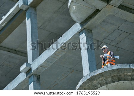 An architect in a protective suit at the height of a construction site with a drawing and a walkie talkie in his hands
