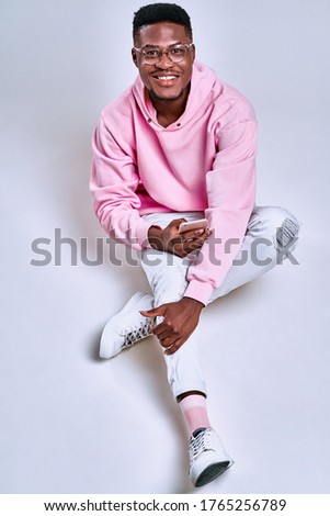Full length of a cheerful young man sitting with legs crossed isolated over white background, using mobile phone. Vertical photo