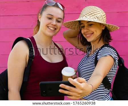 two young girls tourists couple with hat smartphone and coffee cup taking a selfie smiling with pink background