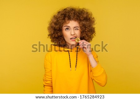 Let's try cryptocurrency! Portrait of curly-haired young woman in urban style hoodie biting golden bitcoin, tasting btc coin on her tooth, blockchain concept. studio shot isolated on yellow background Royalty-Free Stock Photo #1765284923