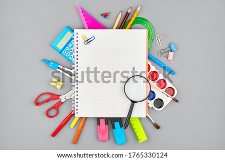 Colored stationery, watercolors, compasses, markers, pencils, and a calculator are covered with an open notebook on a gray wooden table. The view from the top. Space for text. Preparation for school.