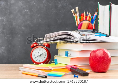 Textbooks, pencils in a stand, a magnifying glass, an alarm clock, a calculator and stationery on a wooden table against a blackboard. Concept of preparation for the beginning of the new academic year