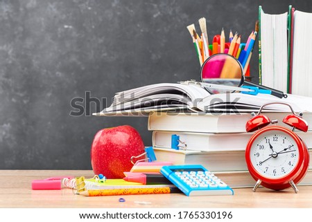 Textbooks, pencils in a stand, a magnifying glass, an alarm clock, a calculator and stationery on a wooden table against a blackboard. Concept of preparation for the beginning of the new academic year