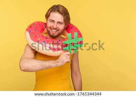 Happy funny tourist guy in undershirt standing with pink donut rubber ring around head and holding hashtag symbol, hash sign of viral web idea, blogging. studio shot isolated on yellow background
