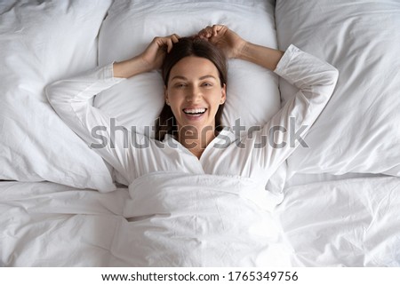 Top view portrait of smiling millennial woman lying relaxing on comfortable white bed on fluffy pillow, happy young female rest wake up in the morning after good sleep in cozy home or hotel bedroom