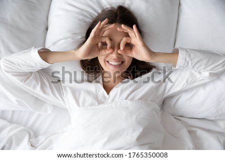 Top view portrait of happy optimistic woman lying in white cozy bed wake up in the morning, smiling young female awaken in comfortable home or hotel bedroom make funny gesture welcome new day