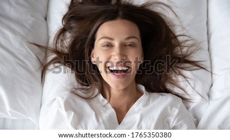 Top view portrait of happy millennial female rest wake up on fluffy pillow in white bed look at camera posing, smiling young woman awaken in cozy bedroom in the morning after good night sleep