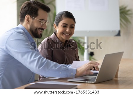 Smiling diverse colleagues working on project together, happy Caucasian businessman and Indian businesswoman sitting at desk, looking at laptop screen, discussing strategy, sharing ideas Royalty-Free Stock Photo #1765351433