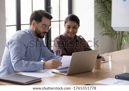 Confident businessman mentor teaching smiling Indian intern, reading documents, diverse colleagues sitting at desk with laptop, working on project together, manager consulting client at meeting Royalty-Free Stock Photo #1765351778
