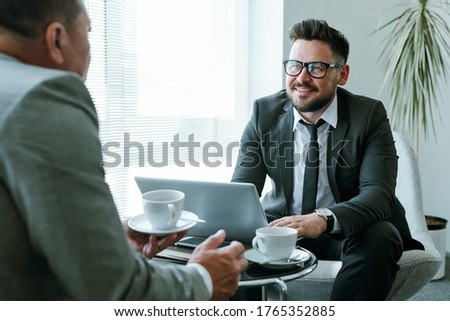 Happy young businessman in suit and eyeglasses looking at his business partner with smile during discussion of new strategies at meeting