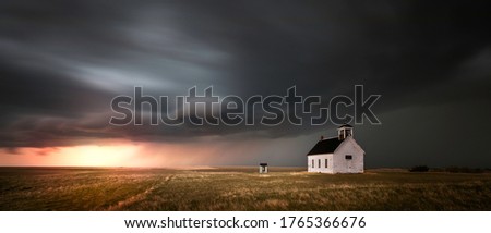 Old church in the rural countryside with a sever storm at sunset. There is an outhouse visible in the scene as well as a green and yellow grass meadow. Royalty-Free Stock Photo #1765366676