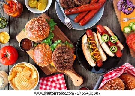 Summer BBQ food table scene with hot dog and hamburger buffet. Top view over a dark wood background.