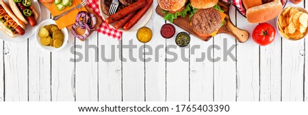 Summer BBQ food table scene with hot dog and hamburger buffet. Above view top border over a white wood banner background. Copy space.