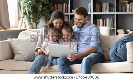 Happy parents with two little daughters using laptop together, smiling mother and father with kids looking at computer screen, watching cartoons, making video call, shopping online, sitting on couch
