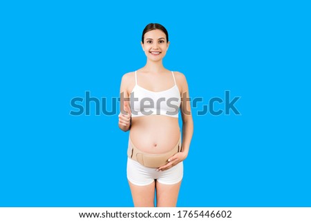 Portrait of young pregnant woman in underwear wearing bandage on her belly and showing thumb up sign at blue background with copy space. Orthopedic abdominal support belt concept.