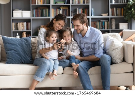 Happy young parents with two adorable little daughters using phone, looking at screen, sitting on cozy couch at home, family making video call, shopping or chatting online, enjoying leisure time