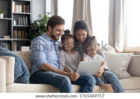 Happy young family with two little daughters using laptop together, smiling mother and father with adorable preschool girls looking at screen, watching cartoons, shopping online, making video call