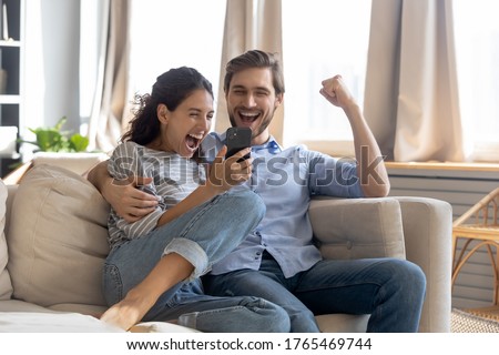 Overjoyed surprised young couple looking at phone screen, reading message with good news, excited woman and man celebrating success, online lottery win, showing yes gesture, sitting on couch Royalty-Free Stock Photo #1765469744