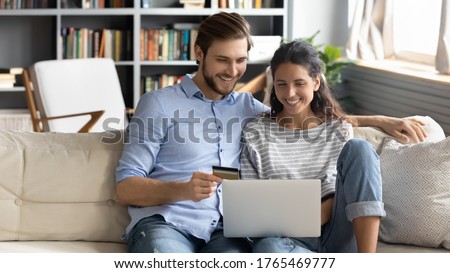 Happy young couple shopping together, customers paying by credit card online, sitting on cozy couch in living room at home, smiling man and woman making internet payment, looking at laptop screen Royalty-Free Stock Photo #1765469777