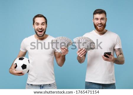 Excited men guys friends in white t-shirt isolated on pastel blue background. Sport leisure lifestyle concept. Cheer up support favorite team with soccer ball hold mobile phone, fan of cash money