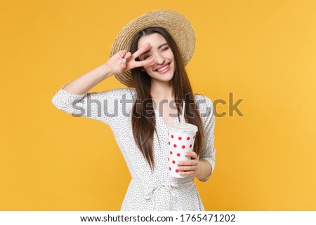 Funny young brunette woman girl in white dress hat posing isolated on yellow background studio. People lifestyle concept. Mock up copy space. Hold plastic cup of soda or cola, showing victory sign