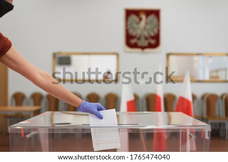 Woman with face mask and gloves, due to pandemic Covid-19, throwing a card with a vote
to the ballot box during  elections. In the backround polish emblem and flag