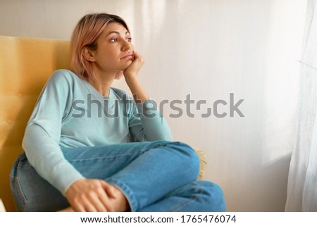 Portrait of beautiful young woman in casual clothes sitting in armchair with hand under chin having sad facial expressing, being bored, spending home alone, copy space for your information
