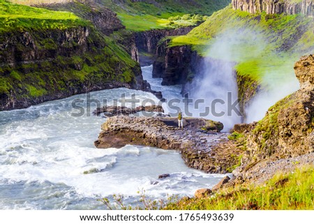 Iceland. Gullfoss "Golden Falls" - the waterfall fed by glacial water. Woman - photographer with green backpack takes pictures of burrowing waterfall. The concept of extreme and photo tourism