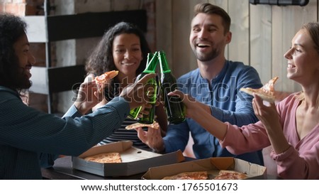 Multi racial excited friends eating pizza hold bottles with alcohol beverage clinking with beer, celebrate success, life event in cafe, relaxing together in public place. Friendship, lifestyle concept