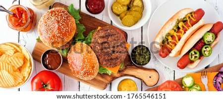 Summer BBQ food table scene with hot dog and hamburger buffet. Top down view banner over a white wood background.