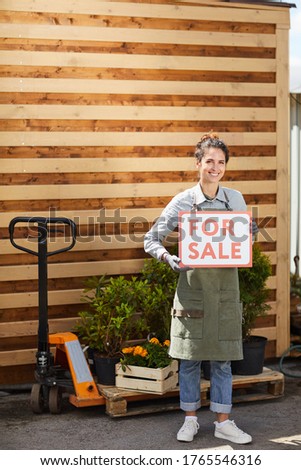 Vertical full length portrait of smiling female worker holding red For Sale sign while standing by plantation outdoors, copy space