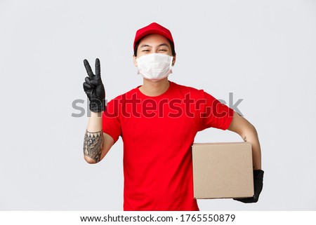Concept of delivery, coronavirus pandemic. Cheerful smiling asian courier in red cap, t-shirt, show peace sign, holding parcel, wear medical mask and gloves for client safety, deliver package