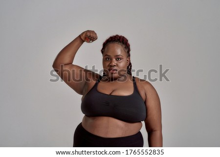 Plump, plus size african american woman in sportswear looking at camera, showing strong arm in studio over grey background. Concept of sport, body positive, equality. Horizontal shot. Front view