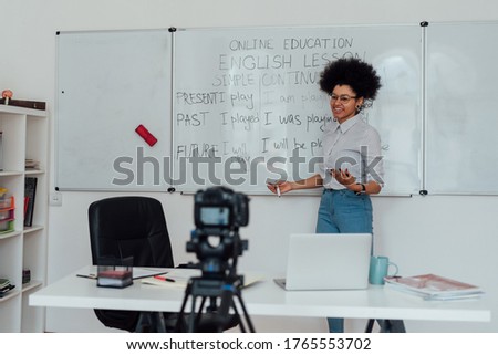 Giving online class. Young afro american female English teacher standing near whiteboard and smiling, explaining rules of English grammar online. Main focus on woman. E-learning, distance education Royalty-Free Stock Photo #1765553702