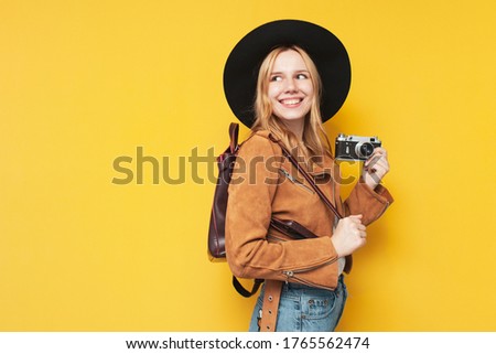 young girl tourist in autumn clothes with a camera and a backpack on a yellow isolated background, travel concept
