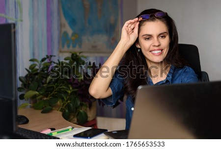 Smiling young woman in denim shirt works online with a laptop in the office at desk or at home, looks to the screen and holds glasses on her head with hand. World map in the background. Blur world map