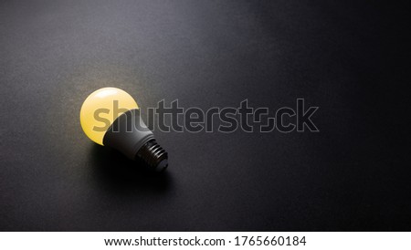 Bright yellow light bulb on black texture background, office desk table from top view, copy space.