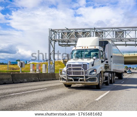 Day cab big rig semi truck with spoiler on the roof and grille guard transporting frozen commercial cargo in refrigerated semi trailer running on the wide highway under the road signage trusses Royalty-Free Stock Photo #1765681682