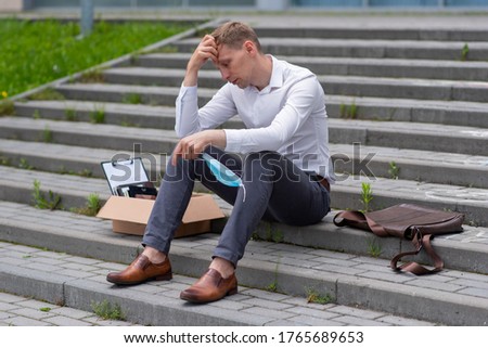Dismissal of an employee due to a coronavirus epidemic. A man sits on the steps. Next to him is his stationery. He has a medical mask in his hand.
