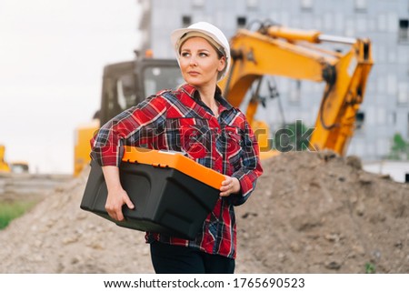 Woman in construction clothes with a white helmet in a plaid shirt on the background of an excavator. Engineer concept at a construction site next to pit excavation equipment