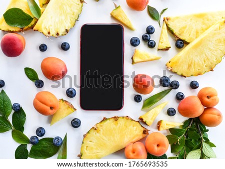 Blank screen smartphone surrounded with berries, apricots, pineapple slices mint and leaves on white table. Top view with free text copy space