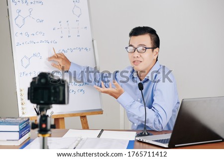 Young Asian chemistry teacher explaining formulas on whiteboard when hosting online class for school students