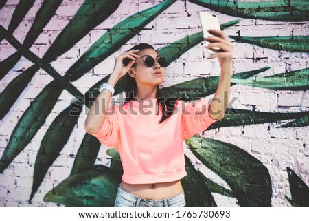 Attractive millennial content maker shooting influence video vlog near photo zone enjoying networking lifestyle, Asian hipster girl in sunglasses smiling at smartphone camera for clicking selfie image
