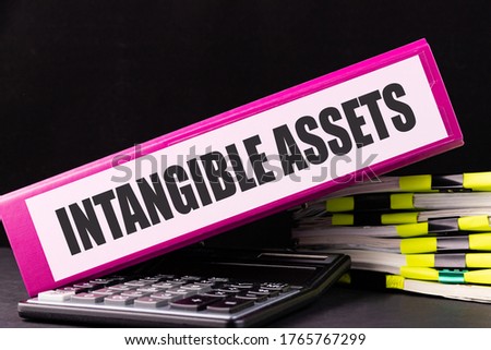 INTANGIBLE ASSETS text is written on a folder lying on a stack of papers on an office desk. Business concept. Royalty-Free Stock Photo #1765767299