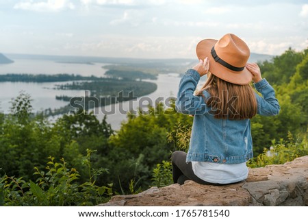 A little girl a child in a denim jacket sits on the edge of a cliff holding her hat in her hands and looks at the amazing natural picture with mountains and a river